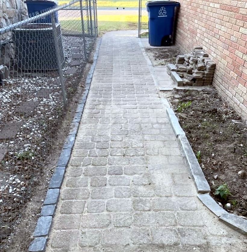A finished paver walkway down the side of a brick house, with a dark border on both edges